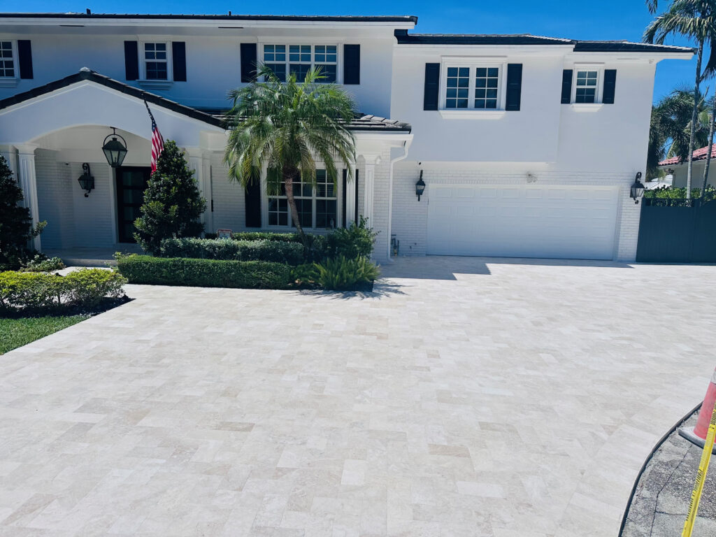 Front view of a white two-story home with dark shutters, palm trees, and a large tiled driveway under a bright blue sky.